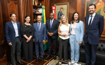 Jorginho Mello recebe embaixador de Ruanda em Florianópolis Fotografia em grupo no gabinete do Governador de Santa Catarina, registrando o encontro diplomático entre o estado e Ruanda. Sete autoridades estão de pé, sorrindo para a câmera, sobre um piso de taco de madeira xadrez, em frente a paredes de painéis de madeira ricamente entalhados. No centro, o Governador Jorginho Mello, vestindo a faixa estadual sobre um terno azul, e o Embaixador de Ruanda no Brasil, Lawrence Manzi, em um terno escuro central com gravata azul, seguram a posição central, simbolizando o acordo de cooperação técnica em educação e agronegócio. Eles são ladeados por secretários estaduais (incluindo Vânia Franco, Luciane Ceretta, Indianara Toreti) e representantes da InvestSC e da embaixada, todos formalmente vestidos. Ao fundo, as bandeiras de Santa Catarina e do Brasil estão proeminentes, ao lado de estantes com troféus e um ícone religioso bizantino, em um ambiente formal, sofisticado e de aproximação internacional.