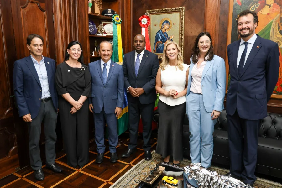 Fotografia em grupo no gabinete do Governador de Santa Catarina, registrando o encontro diplomático entre o estado e Ruanda. Sete autoridades estão de pé, sorrindo para a câmera, sobre um piso de taco de madeira xadrez, em frente a paredes de painéis de madeira ricamente entalhados. No centro, o Governador Jorginho Mello, vestindo a faixa estadual sobre um terno azul, e o Embaixador de Ruanda no Brasil, Lawrence Manzi, em um terno escuro central com gravata azul, seguram a posição central, simbolizando o acordo de cooperação técnica em educação e agronegócio. Eles são ladeados por secretários estaduais (incluindo Vânia Franco, Luciane Ceretta, Indianara Toreti) e representantes da InvestSC e da embaixada, todos formalmente vestidos. Ao fundo, as bandeiras de Santa Catarina e do Brasil estão proeminentes, ao lado de estantes com troféus e um ícone religioso bizantino, em um ambiente formal, sofisticado e de aproximação internacional.