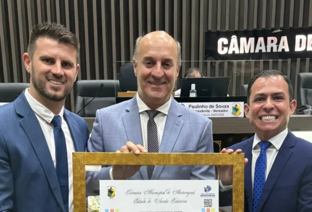 Jos&eacute; Milton Scheffer, em terno cinza, sorri e segura o quadro de Cidad&atilde;o Honor&aacute;rio de Ararangu&aacute;, ladeado por dois vereadores da C&acirc;mara de Vereadores de Ararangu&aacute;. O quadro tem o bras&atilde;o da C&acirc;mara e o texto 'Estado de Santa Catarina'. No fundo, placas de identifica&ccedil;&atilde;o de vereadores.