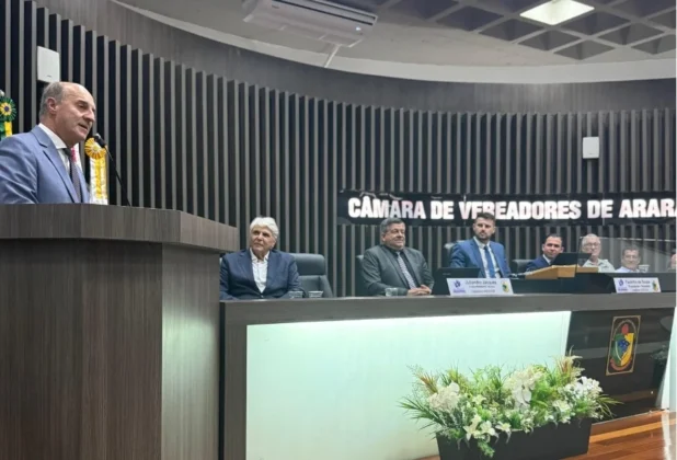 Jos&eacute; Milton Scheffer, em terno cinza, discursa no p&uacute;lpito da C&acirc;mara de Vereadores de Ararangu&aacute;. Outros vereadores est&atilde;o sentados atr&aacute;s da mesa principal com placas de identifica&ccedil;&atilde;o e arranjos florais. O letreiro 'C&Acirc;MARA DE VEREADORES DE ARARANGU&Aacute;' est&aacute; vis&iacute;vel.
