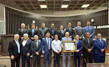 José Milton Scheffer recebe título de cidadão em Araranguá Uma foto de grupo com quatorze homens em trajes formais na Câmara Municipal de Araranguá. No centro, José Milton Scheffer segura o quadro de Cidadão Honorário, cercado por vereadores e autoridades. O letreiro 'CÂMARA DE ARARANGUÁ' está visível ao fundo.