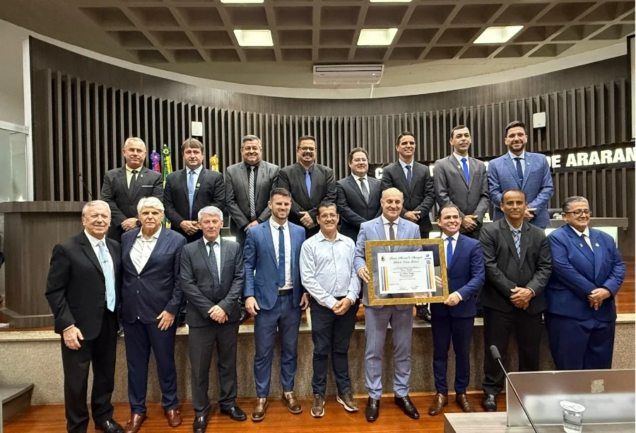 Uma foto de grupo com quatorze homens em trajes formais na Câmara Municipal de Araranguá. No centro, José Milton Scheffer segura o quadro de Cidadão Honorário, cercado por vereadores e autoridades. O letreiro 'CÂMARA DE ARARANGUÁ' está visível ao fundo.