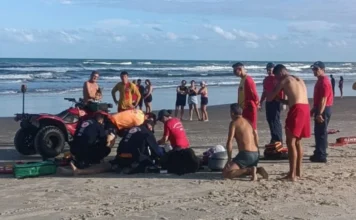 Jovem de 18 anos morre afogado em Passo de Torres Imagem capturada na areia da Praia Pérola, em Passo de Torres, mostrando a mobilização de socorristas em torno de uma vítima de afogamento. No centro, profissionais do Corpo de Bombeiros e do SAER, vestindo uniformes vermelhos e azuis escuros, estão ajoelhados realizando manobras de reanimação. À esquerda, um quadriciclo vermelho de salvamento está estacionado. Ao fundo, é possível ver a linha do mar com ondas moderadas e alguns banhistas observando a operação sob um céu com poucas nuvens.