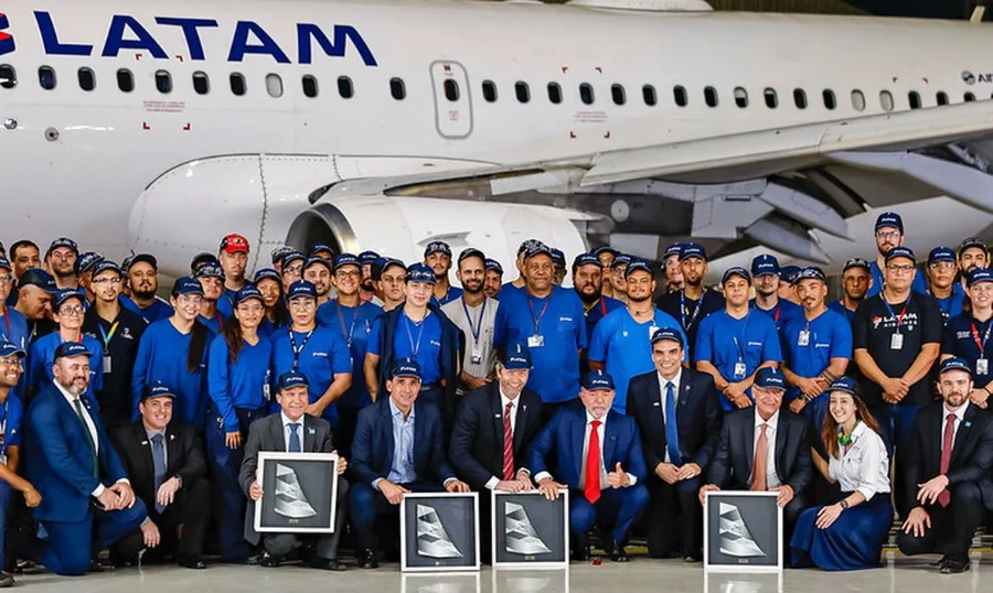Latam incorpora 24 aviões da Embraer para voos regionais Foto de grupo oficial no hangar MRO em São Carlos, SP, celebrando a entrega dos primeiros jatos Embraer E195-E2 para a Latam. O presidente Lula e o vice-presidente Alckmin estão posicionados na frente com executivos da Latam e da Embraer, rodeados por centenas de funcionários uniformizados. Quatro executivos seguram placas com o design da cauda do avião E2.
