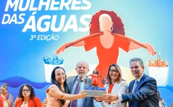 Lula garante manutenção do seguro-defeso para pescadores Presidente Luiz Inácio Lula da Silva e primeira-dama Janja da Silva, junto com autoridades e premiada, segurando o troféu da 3ª Edição do Prêmio Mulheres das Águas. Ao fundo, grande banner ilustrado do evento com texto e a figura de uma mulher segurando baldes com peixes e caranguejos, representando o reconhecimento de mulheres na pesca artesanal e aquicultura no Brasil.