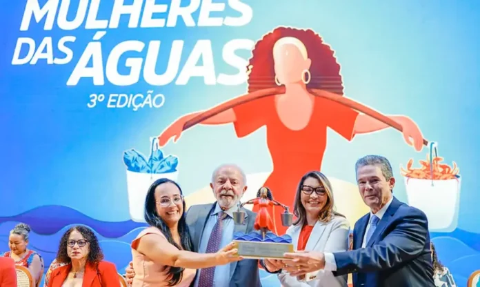 Lula garante manutenção do seguro-defeso para pescadores Presidente Luiz Inácio Lula da Silva e primeira-dama Janja da Silva, junto com autoridades e premiada, segurando o troféu da 3ª Edição do Prêmio Mulheres das Águas. Ao fundo, grande banner ilustrado do evento com texto e a figura de uma mulher segurando baldes com peixes e caranguejos, representando o reconhecimento de mulheres na pesca artesanal e aquicultura no Brasil.