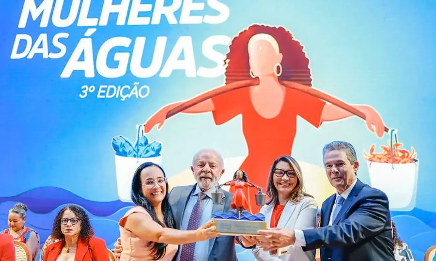 Presidente Luiz Inácio Lula da Silva e primeira-dama Janja da Silva, junto com autoridades e premiada, segurando o troféu da 3ª Edição do Prêmio Mulheres das Águas. Ao fundo, grande banner ilustrado do evento com texto e a figura de uma mulher segurando baldes com peixes e caranguejos, representando o reconhecimento de mulheres na pesca artesanal e aquicultura no Brasil.