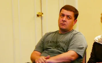 Lulinha admite viagem paga por lobista preso do INSS Fotografia colorida em plano médio de Fábio Luís Lula da Silva, homem de meia-idade, com cabelos curtos e grisalhos, vestindo um paletó azul-marinho sobre uma camisa social branca de gola aberta. Ele está caminhando em um ambiente externo desfocado, com uma expressão séria e olhando ligeiramente para a esquerda. A imagem é um registro jornalístico.