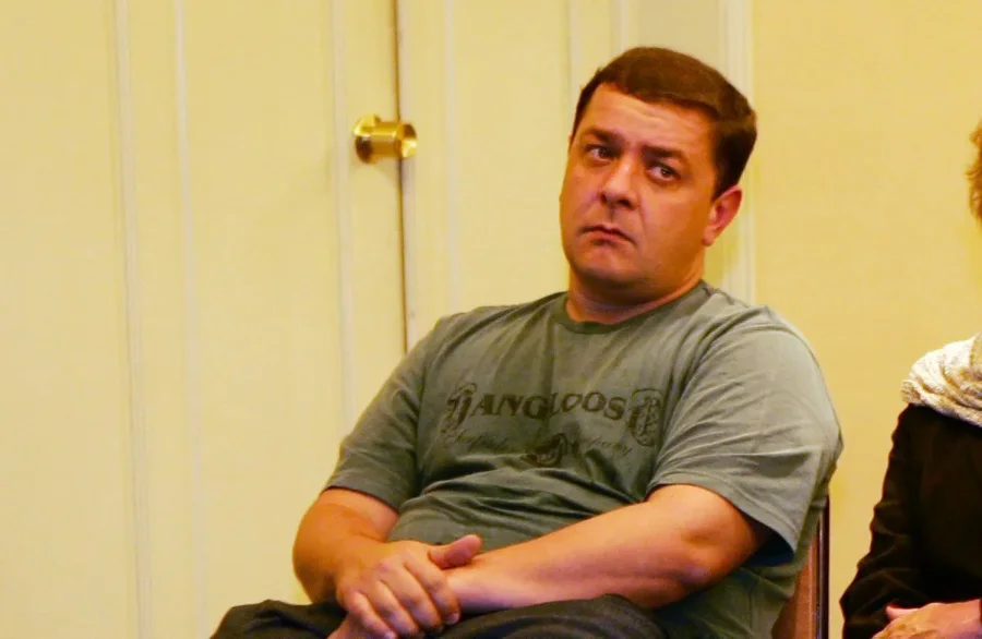 Lulinha admite viagem paga por lobista preso do INSS Fotografia colorida em plano médio de Fábio Luís Lula da Silva, homem de meia-idade, com cabelos curtos e grisalhos, vestindo um paletó azul-marinho sobre uma camisa social branca de gola aberta. Ele está caminhando em um ambiente externo desfocado, com uma expressão séria e olhando ligeiramente para a esquerda. A imagem é um registro jornalístico.