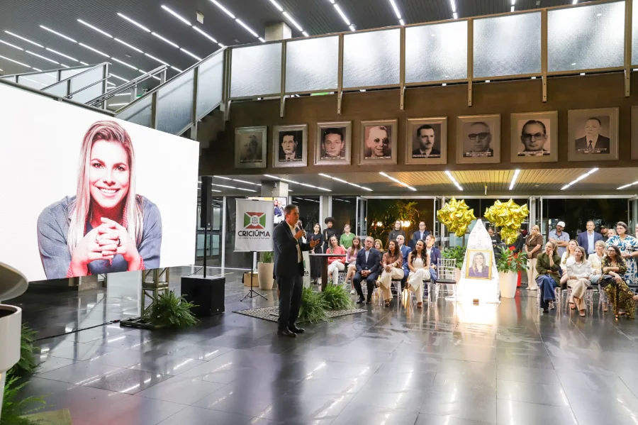 Fotografia colorida da cerimônia oficial de entrega da Medalha Thati Teixeira, no hall do Paço Municipal Marcos Rovaris, em Criciúma. O amplo espaço moderno tem pisos de ardósia escura e luminárias lineares no teto. No centro-esquerda, um homem de terno preto e microfone na mão fala em pé num pequeno palco improvisado, ladeado por um banner com o brasão da Prefeitura de Criciúma e plantas em vasos. Atrás dele, na parede, uma grande tela LED exibe o retrato sorridente de uma mulher com cabelos loiros e mãos entrelaçadas. Acima da tela, há uma galeria com 10 retratos emoldurados de ex-prefeitos. Uma audiência de cerca de 30 pessoas, homens e mulheres, assiste sentada em cadeiras transparentes e de madeira à direita. No primeiro plano à direita, há um altar iluminado e decorado com flores brancas, velas e uma foto emoldurada da mesma mulher, ladeado por dois grandes cachos de balões dourados em forma de estrela. Grandes janelas de vidro e folhagens são visíveis ao fundo, sugerindo um jardim externo.