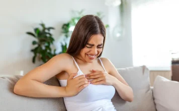 Médica destaca sinais de infarto em mulheres Uma mulher jovem com cabelos castanhos longos está sentada em um sofá bege, vestindo uma regata branca. Ela tem uma expressão de dor intensa no rosto e está apertando a área central e esquerda do peito com as duas mãos. O fundo é uma sala de estar clara e desfocada, com plantas em vasos. A imagem ilustra visualmente a dor no peito, um sintoma de problemas cardíacos em mulheres, alinhando-se com o alerta da Dra. Araceli Tomaz sobre a saúde cardiovascular feminina.