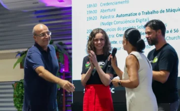 Meetup Startup SC reúne ecossistema de inovação no CRIO Foto tirada no palco do CRIO (Unesc) durante o evento Meetup Startup SC. Quatro pessoas estão presentes, interagindo com o público e entre si. Ao fundo, um grande projetor exibe o cronograma do evento, com os horários de credenciamento, abertura e o título da palestra de Fábio Nudge sobre automatização do trabalho de máquinas. À esquerda, um homem calvo com óculos e camisa polo estende a mão para um cumprimento. No centro-esquerda, Elenice Engel, gerente de Inovação da Unesc, vestindo uma camisa preta com logo da Unesc e uma saia vermelha longa, aplaude em destaque. No centro-direita, uma mulher com vestido branco segura um microfone, dirigindo-se ao homem da esquerda. À direita, um homem de barba e camisa preta com um logo assiste. A cena capta a atmosfera de troca e networking promovida pelo encontro entre a universidade, o mercado e instituições de fomento.