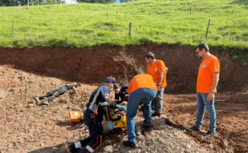 Meleiro faz simulado de desastre com apoio do Estado A imagem mostra equipes de resgate trabalhando em um cenário de simulado de deslizamento de terra em Meleiro. Em um terreno de terra vermelha escavada, profissionais de uniformes azul-marinho com faixas refletivas e camisetas laranjas, representando o Corpo de Bombeiros, SAMU e equipes de apoio municipal, prestam socorro a duas vítimas simuladas. Uma pessoa está deitada no chão à esquerda, suja de terra. Outra pessoa está sendo imobilizada em uma maca de resgate amarela no centro por socorristas agachados. Dois outros homens de camisetas laranja estão em pé ao lado, observando e apoiando a operação. Ao fundo, vê-se um barranco de terra cortado e uma colina gramada com cercas.
