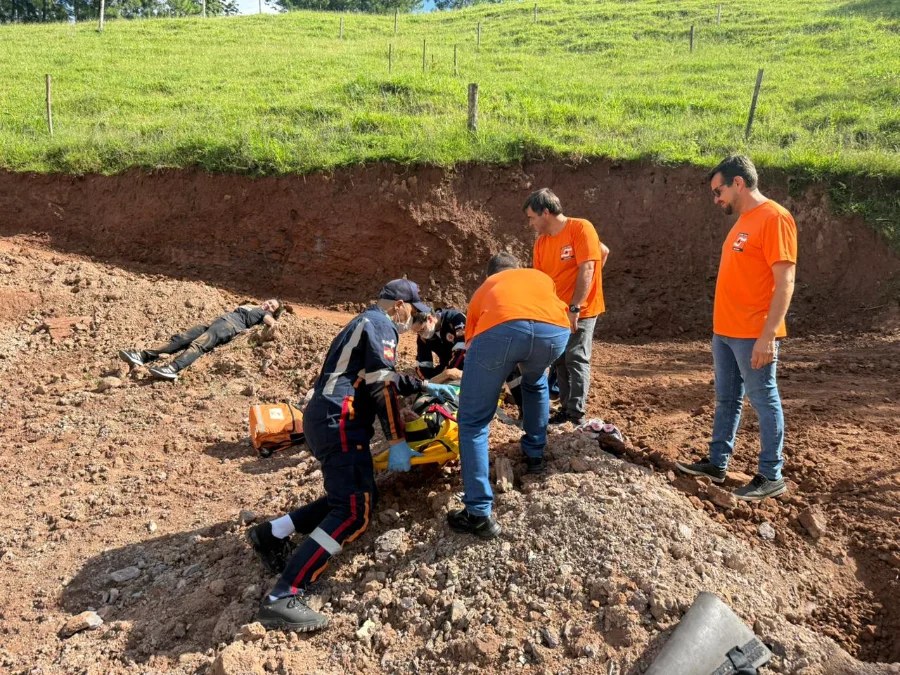 Meleiro faz simulado de desastre com apoio do Estado A imagem mostra equipes de resgate trabalhando em um cenário de simulado de deslizamento de terra em Meleiro. Em um terreno de terra vermelha escavada, profissionais de uniformes azul-marinho com faixas refletivas e camisetas laranjas, representando o Corpo de Bombeiros, SAMU e equipes de apoio municipal, prestam socorro a duas vítimas simuladas. Uma pessoa está deitada no chão à esquerda, suja de terra. Outra pessoa está sendo imobilizada em uma maca de resgate amarela no centro por socorristas agachados. Dois outros homens de camisetas laranja estão em pé ao lado, observando e apoiando a operação. Ao fundo, vê-se um barranco de terra cortado e uma colina gramada com cercas.
