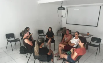 Meleiro lança programa de apoio a mães de crianças autistas Fotografia em plano médio mostra sete mulheres sentadas em cadeiras pretas dispostas em círculo, em uma sala de paredes brancas e piso claro. Elas participam de uma roda de conversa em um ambiente de reunião. Ao fundo, há uma mesa com papéis e uma tela de projeção branca que exibe a mensagem "SEM SINAL".
