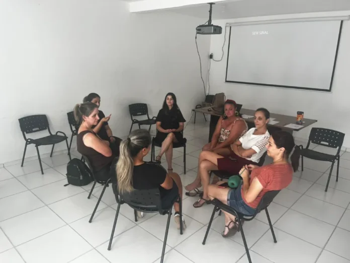 Meleiro lança programa de apoio a mães de crianças autistas Fotografia em plano médio mostra sete mulheres sentadas em cadeiras pretas dispostas em círculo, em uma sala de paredes brancas e piso claro. Elas participam de uma roda de conversa em um ambiente de reunião. Ao fundo, há uma mesa com papéis e uma tela de projeção branca que exibe a mensagem "SEM SINAL".