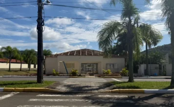 Meleiro reforma Centro Multiuso em parceria com o Estado Fachada do Centro Multiuso João Macarini em Meleiro, SC, em processo de reforma, com o telhado removido e a estrutura de madeira exposta para recuperação. Imagem de obra pública com entulho de construção e palmeiras, vista da rua com faixas de pedestres.