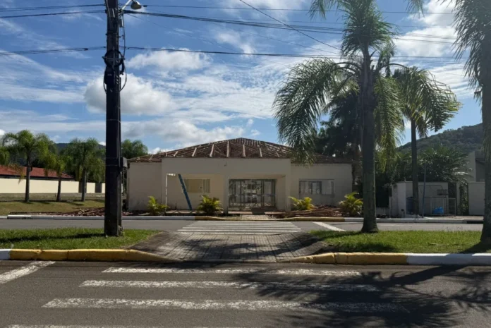 Meleiro reforma Centro Multiuso em parceria com o Estado Fachada do Centro Multiuso João Macarini em Meleiro, SC, em processo de reforma, com o telhado removido e a estrutura de madeira exposta para recuperação. Imagem de obra pública com entulho de construção e palmeiras, vista da rua com faixas de pedestres.