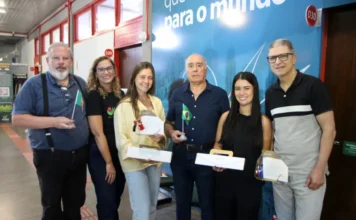 Mestres da Unesc recebem diploma europeu de engenharia Fotografia em plano médio mostra seis pessoas em pé, posando para foto em um ambiente interno da Unesc. No centro, o professor Manuel Ribeiro, um homem senhor de camisa azul escura, segura uma pequena bandeira do Brasil. Ele está ladeado pelas mestres Eduarda Fraga Olivo e Mariana de Souza Pereira, que sorriem e seguram seus diplomas e caixas brancas de lembrança. À esquerda, um homem segura uma pequena bandeira de Portugal. Ao fundo, destaca-se um painel azul com a inscrição parcial "para o mundo".