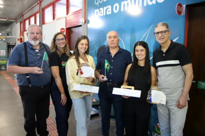 Mestres da Unesc recebem diploma europeu de engenharia Fotografia em plano médio mostra seis pessoas em pé, posando para foto em um ambiente interno da Unesc. No centro, o professor Manuel Ribeiro, um homem senhor de camisa azul escura, segura uma pequena bandeira do Brasil. Ele está ladeado pelas mestres Eduarda Fraga Olivo e Mariana de Souza Pereira, que sorriem e seguram seus diplomas e caixas brancas de lembrança. À esquerda, um homem segura uma pequena bandeira de Portugal. Ao fundo, destaca-se um painel azul com a inscrição parcial "para o mundo".