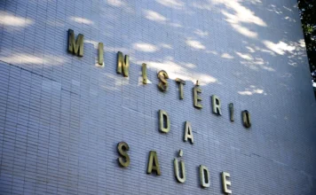 Ministério da Saúde atende mil Yanomamis em força-tarefa Fachada lateral de um edifício revestido com azulejos brancos, onde se lê a inscrição "MINISTÉRIO DA SAÚDE" em letras metálicas douradas. A luz solar incide sobre a parede, criando sombras de folhagens. A imagem simboliza a atuação do órgão federal nas ações de emergência e saúde pública no Território Yanomami.