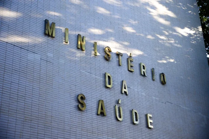 Ministério da Saúde atende mil Yanomamis em força-tarefa Fachada lateral de um edifício revestido com azulejos brancos, onde se lê a inscrição "MINISTÉRIO DA SAÚDE" em letras metálicas douradas. A luz solar incide sobre a parede, criando sombras de folhagens. A imagem simboliza a atuação do órgão federal nas ações de emergência e saúde pública no Território Yanomami.