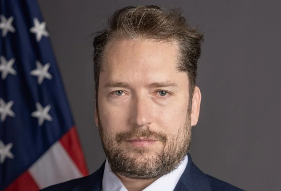 Fotografia em close do rosto de Darren Beattie, um homem branco de cabelos castanhos curtos, olhos claros e barba curta. Ao fundo, do lado esquerdo, aparece parte de uma bandeira dos Estados Unidos levemente desfocada.