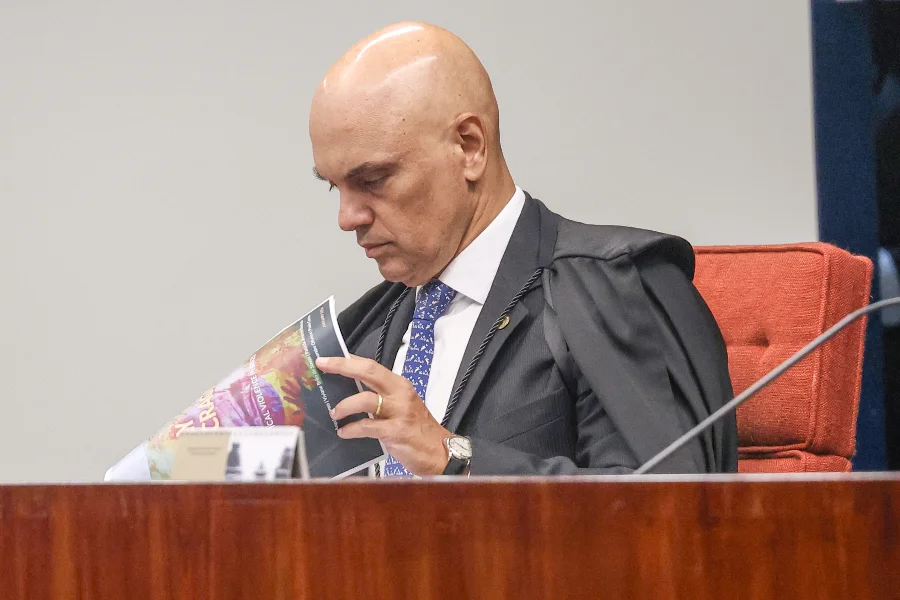 Fotografia de perfil do ministro Alexandre de Moraes , do Supremo Tribunal Federal (STF), sentado em uma cadeira de encosto vermelho. Ele veste a toga preta tradicional da Corte sobre terno e gravata azul estampada. O ministro está com a cabeça levemente inclinada, folheando e lendo atentamente um documento impresso com elementos coloridos sobre uma bancada de madeira.