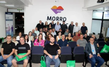 Morro da Fumaça planeja setor cerâmico para os próximos 30 anos Fotografia registra o grupo de empresários, pesquisadores e autoridades reunidos em um auditório durante a apresentação do planejamento estratégico do APL de Cerâmica Vermelha, em Morro da Fumaça. Cerca de 30 pessoas aparecem na imagem; as que estão na primeira fila aparecem sentadas em poltronas azuis segurando sacolas ecológicas verdes com o logotipo da Unesc, enquanto o restante do grupo permanece em pé ao fundo, posicionado abaixo da identidade visual oficial do município.