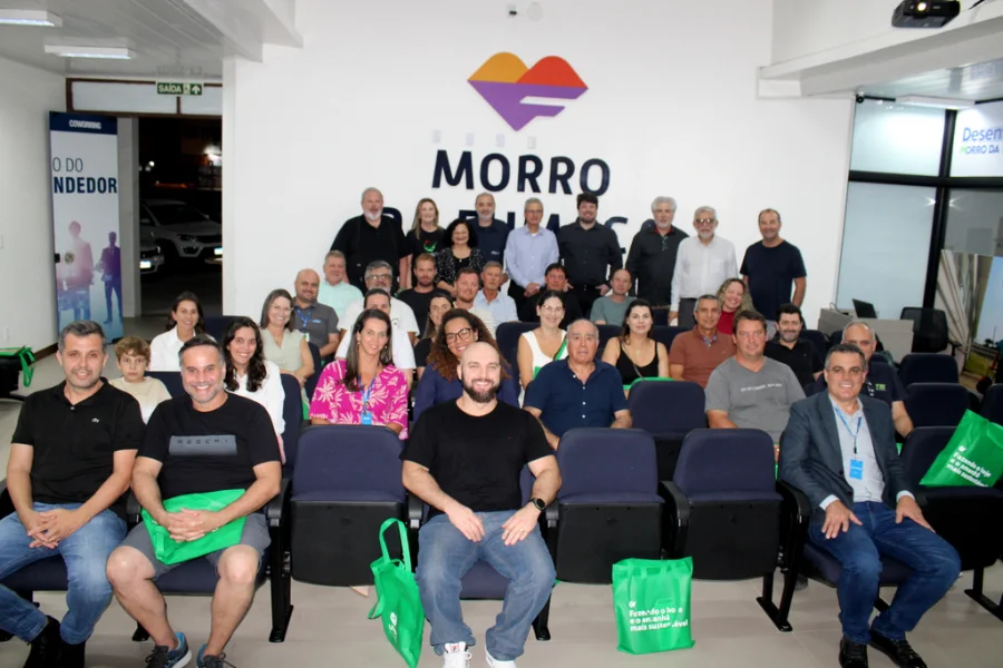 Fotografia registra o grupo de empresários, pesquisadores e autoridades reunidos em um auditório durante a apresentação do planejamento estratégico do APL de Cerâmica Vermelha, em Morro da Fumaça. Cerca de 30 pessoas aparecem na imagem; as que estão na primeira fila aparecem sentadas em poltronas azuis segurando sacolas ecológicas verdes com o logotipo da Unesc, enquanto o restante do grupo permanece em pé ao fundo, posicionado abaixo da identidade visual oficial do município.