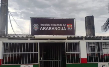 Mudança nas visitas no Presídio de Araranguá por superlotação Fotografia da fachada do Presídio Regional de Araranguá. No centro da imagem, destaca-se uma placa marrom com os dizeres "Presídio Regional de Araranguá" em letras brancas, ladeada pelos brasões da Polícia Penal e da Secretaria de Administração Prisional (SAP). A estrutura da entrada apresenta grades de segurança e paredes pintadas com faixas nas cores branca, vermelha e verde, sob um céu nublado.
