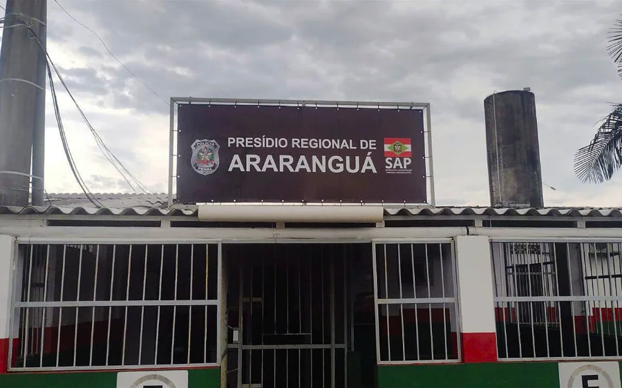 Fotografia da fachada do Presídio Regional de Araranguá. No centro da imagem, destaca-se uma placa marrom com os dizeres 