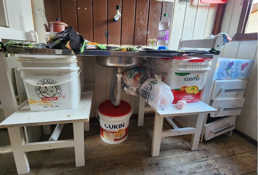 Pia de cozinha improvisada apoiada sobre cadeiras e baldes plásticos na residência de Dona Neusa, moradora do bairro São Luiz em Sombrio. A imagem detalha a precariedade das instalações e a necessidade urgente de doação de um balcão de pia e móveis básicos.
