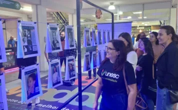 Mulheres na tecnologia são tema de exposição imersiva Fotografia da exposição "Conexões que Inspiram" em um saguão universitário. No centro, um suporte metálico sustenta fios de aço onde vários cartazes estão pendurados por grampos, formando um varal. Cada cartaz apresenta a foto de uma mulher pioneira na tecnologia, como Hedy Lamarr e a brasileira Claudia Bauzer Medeiros, acompanhada de um QR Code para acesso ao conteúdo digital. À direita, um grupo de pessoas, majoritariamente mulheres, observa atentamente a mostra. No primeiro plano, uma mulher veste uma camiseta preta com o logotipo da Unesc e sorri enquanto aprecia os cartazes.