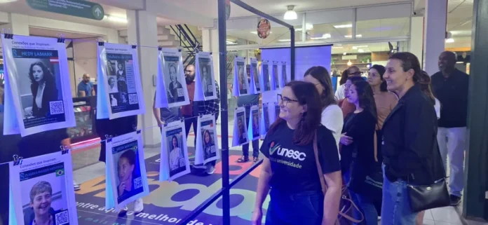 Mulheres na tecnologia são tema de exposição imersiva Fotografia da exposição "Conexões que Inspiram" em um saguão universitário. No centro, um suporte metálico sustenta fios de aço onde vários cartazes estão pendurados por grampos, formando um varal. Cada cartaz apresenta a foto de uma mulher pioneira na tecnologia, como Hedy Lamarr e a brasileira Claudia Bauzer Medeiros, acompanhada de um QR Code para acesso ao conteúdo digital. À direita, um grupo de pessoas, majoritariamente mulheres, observa atentamente a mostra. No primeiro plano, uma mulher veste uma camiseta preta com o logotipo da Unesc e sorri enquanto aprecia os cartazes.