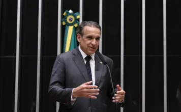 Câmara aprova cobrança de multa retroativa para ex-dono Uma foto colorida mostra o deputado federal Hildo Rocha de pé, atrás de um púlpito com um microfone, na Câmara dos Deputados. Ele veste um terno cinza escuro, camisa branca e gravata listrada de azul marinho. Suas mãos estão levantadas e gesticulando para a frente, sugerindo que ele está discursando. O fundo é composto por painéis pretos com listras verticais prateadas. À esquerda de Rocha, a bandeira do Brasil está pendurada dobrada em um suporte. Sua expressão é séria e engajada.