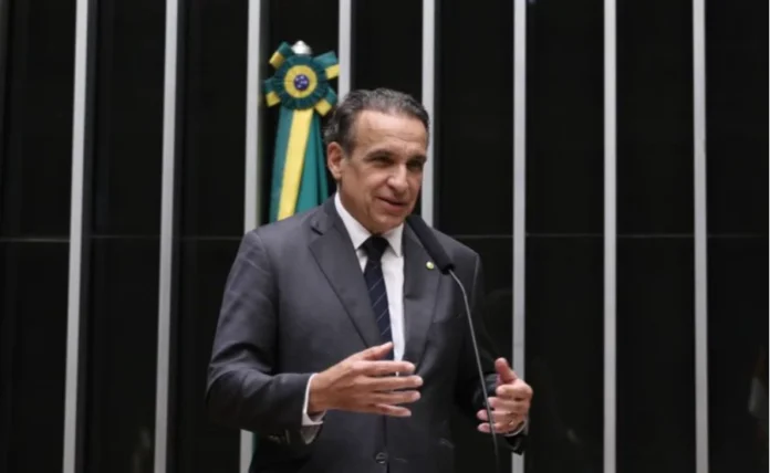 Multas antigas não recairão mais sobre novo dono de veículo Uma foto colorida mostra o deputado federal Hildo Rocha de pé, atrás de um púlpito com um microfone, na Câmara dos Deputados. Ele veste um terno cinza escuro, camisa branca e gravata listrada de azul marinho. Suas mãos estão levantadas e gesticulando para a frente, sugerindo que ele está discursando. O fundo é composto por painéis pretos com listras verticais prateadas. À esquerda de Rocha, a bandeira do Brasil está pendurada dobrada em um suporte. Sua expressão é séria e engajada.