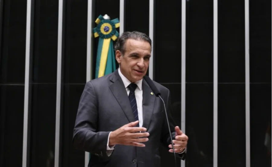 Uma foto colorida mostra o deputado federal Hildo Rocha de pé, atrás de um púlpito com um microfone, na Câmara dos Deputados. Ele veste um terno cinza escuro, camisa branca e gravata listrada de azul marinho. Suas mãos estão levantadas e gesticulando para a frente, sugerindo que ele está discursando. O fundo é composto por painéis pretos com listras verticais prateadas. À esquerda de Rocha, a bandeira do Brasil está pendurada dobrada em um suporte. Sua expressão é séria e engajada.