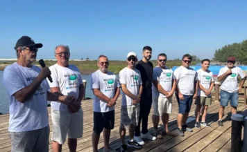 Mutirão em Araranguá recolhe 1,3 tonelada de resíduos Grupo de nove voluntários e autoridades municipais de Araranguá em pé sobre um deck de madeira nas margens do Rio Araranguá para a 5ª edição do Projeto Arco-Íris. O homem da esquerda fala ao microfone. As camisetas brancas mostram o logotipo 'arcoiris' e 'Araranguá'. O fundo exibe a vegetação natural, o rio e as dunas de areia sob um céu azul claro.