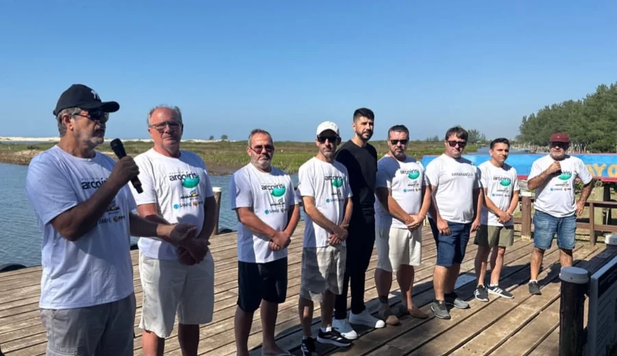 Grupo de nove voluntários e autoridades municipais de Araranguá em pé sobre um deck de madeira nas margens do Rio Araranguá para a 5ª edição do Projeto Arco-Íris. O homem da esquerda fala ao microfone. As camisetas brancas mostram o logotipo 'arcoiris' e 'Araranguá'. O fundo exibe a vegetação natural, o rio e as dunas de areia sob um céu azul claro.
