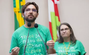 NMO: Alesc debate falta de remédios para doença rara Uma fotografia colorida de nível médio mostra o Dr. Ricardo Pereira Gonçalves, um homem com barba e óculos, no púlpito de oradores, falando ao microfone com as mãos gesticulando para ênfase. Ele veste uma camiseta verde vibrante com o texto impresso: "Você já falou da NMO para alguém hoje?". À sua direita, em segundo plano, está a jornalista Márcia Denardin, também com óculos e a mesma camiseta verde, com um colar de girassóis visível. O fundo é de uma sessão legislativa da ALESC (Assembleia Legislativa de Santa Catarina), com bandeiras estaduais desfocadas e microfones duplos. A iluminação é clara e a expressão do orador é séria e engajada, refletindo a importância do tema durante a campanha Março Verde de conscientização sobre a NMO.