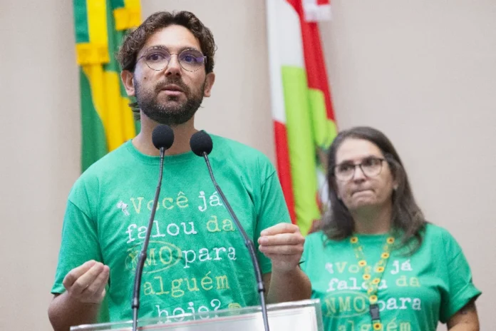 NMO Alesc debate falta de remédios para doença rara Uma fotografia colorida de nível médio mostra o Dr. Ricardo Pereira Gonçalves, um homem com barba e óculos, no púlpito de oradores, falando ao microfone com as mãos gesticulando para ênfase. Ele veste uma camiseta verde vibrante com o texto impresso: "Você já falou da NMO para alguém hoje?". À sua direita, em segundo plano, está a jornalista Márcia Denardin, também com óculos e a mesma camiseta verde, com um colar de girassóis visível. O fundo é de uma sessão legislativa da ALESC (Assembleia Legislativa de Santa Catarina), com bandeiras estaduais desfocadas e microfones duplos. A iluminação é clara e a expressão do orador é séria e engajada, refletindo a importância do tema durante a campanha Março Verde de conscientização sobre a NMO.