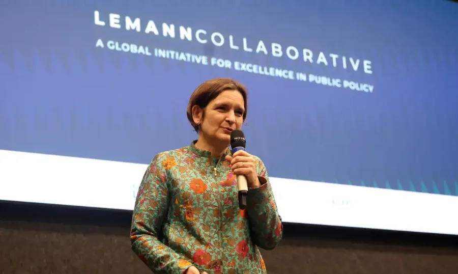 Esther Duflo, economista ganhadora do Prêmio Nobel, palestrando no palco de um evento sobre políticas públicas da Fundação Lemann e Enap em Brasília. Ela segura um microfone e está na frente de uma tela azul com o texto projetado 'LEMANN COLLABORATIVE: A GLOBAL INITIATIVE FOR EXCELLENCE IN PUBLIC POLICY'.