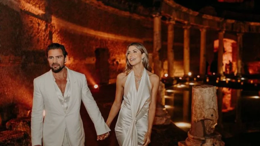 Um homem e uma mulher caminham de mãos dadas em um cenário histórico ao ar livre durante a noite. O homem, à esquerda, veste um terno branco e possui barba aparada. A mulher, à direita, usa um vestido longo de seda branco com decote frente única e um colar de destaque. O fundo apresenta colunas de pedra antigas e ruínas iluminadas por luzes amareladas e quentes, criando uma atmosfera solene.