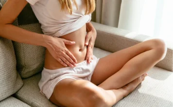 O intestino: nosso segundo cérebro Fotografia em close-up de uma mulher sentada de pernas cruzadas em um sofá texturizado. Ela veste uma camiseta branca dobrada para cima e shorts leves de cor clara. Suas mãos estão apoiadas suavemente sobre a região do abdômen e do intestino, em um gesto de atenção e autocuidado. A luz natural suave entra pela direita, criando uma atmosfera calma. O foco está no tronco e abdômen, evidenciando a conexão entre a pessoa e o centro de sua saúde intestinal.