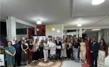 OAB Criciúma lança Comissão de Acadêmicos de Direito na Unesc Uma fotografia interna em plano aberto e amplo captura um grande grupo de aproximadamente 35 pessoas reunidas para uma cerimônia formal no saguão de uma instituição. No centro da imagem, em pé sobre um tapete circular decorativo, três indivíduos seguram e exibem documentos emoldurados (certificados): um homem em terno escuro e gravata, ladeado por duas mulheres com vestidos brancos (uma com estampa floral na saia). Ao redor deles, o restante do grupo, vestido com trajes formais e semi-formais de cores variadas (incluindo verde, vermelho e preto), sorri para a câmera. O ambiente tem pisos de cerâmica, iluminação de teto embutida e painéis decorativos de parede. À direita, é visível um piso tátil vermelho para acessibilidade. A atmosfera é de celebração solene.