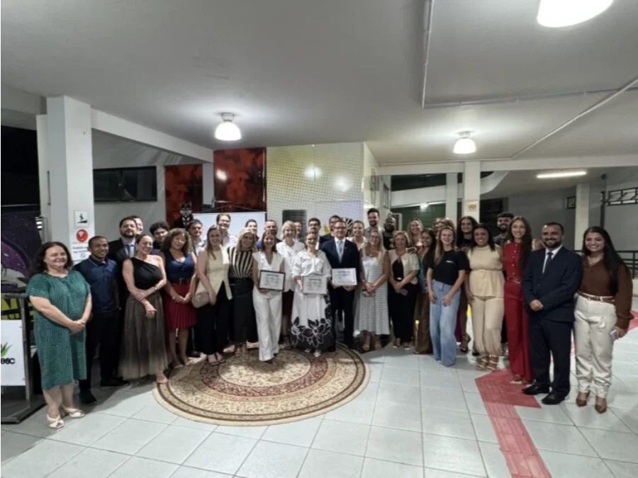 Uma fotografia interna em plano aberto e amplo captura um grande grupo de aproximadamente 35 pessoas reunidas para uma cerimônia formal no saguão de uma instituição. No centro da imagem, em pé sobre um tapete circular decorativo, três indivíduos seguram e exibem documentos emoldurados (certificados): um homem em terno escuro e gravata, ladeado por duas mulheres com vestidos brancos (uma com estampa floral na saia). Ao redor deles, o restante do grupo, vestido com trajes formais e semi-formais de cores variadas (incluindo verde, vermelho e preto), sorri para a câmera. O ambiente tem pisos de cerâmica, iluminação de teto embutida e painéis decorativos de parede. À direita, é visível um piso tátil vermelho para acessibilidade. A atmosfera é de celebração solene.