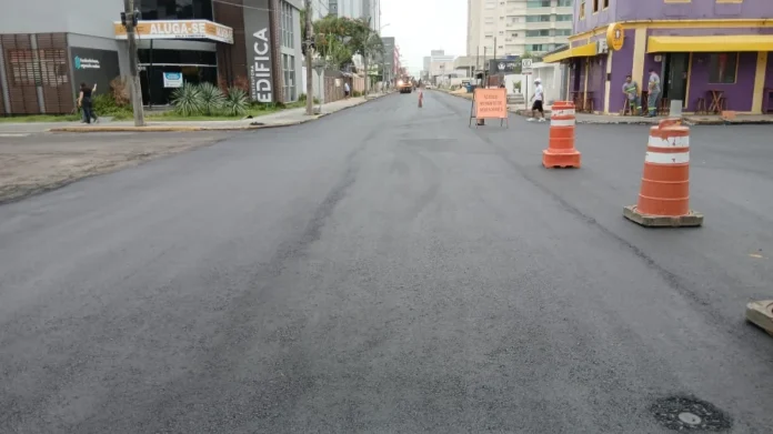 Obra na rua Caetano Lummertz entra em fase final em Araranguá Uma fotografia de nível do solo olhando para o fim de uma rua urbana recém-pavimentada com asfalto escuro e fresco. No primeiro plano, à direita, há dois cones de segurança laranja e brancos. Mais adiante na rua, algumas pessoas são visíveis, incluindo operários de construção com capacetes e coletes de segurança perto de um pequeno maquinário de asfalto. No lado esquerdo, vê-se um prédio moderno de escritórios com placas "ALUGA-SE SALA COMERCIAL" e uma placa vertical "EDIFÍCA". No lado direito, destaca-se um edifício roxo com um toldo amarelo e mesas de pátio ao ar livre. A perspectiva é para baixo ao longo da extensão da rua, mostrando a nova superfície de asfalto.