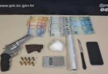Operação policial apreende arma e cocaína em Criciúma Itens apreendidos pela Polícia Militar em Criciúma dispostos sobre uma mesa: um revólver prata, oito munições, uma porção de cocaína, balança de precisão, rolo de plástico filme, faca, celular e diversas notas de Real (notas de 200, 100 e valores menores).