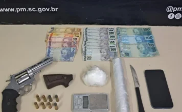 Operação policial apreende arma e cocaína em Criciúma Itens apreendidos pela Polícia Militar em Criciúma dispostos sobre uma mesa: um revólver prata, oito munições, uma porção de cocaína, balança de precisão, rolo de plástico filme, faca, celular e diversas notas de Real (notas de 200, 100 e valores menores).